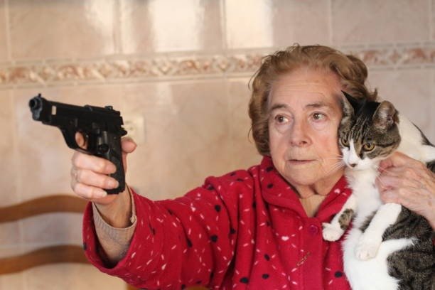 Elderly woman in red heart-printed robe holding a gun with a relaxed tabby cat indoors.