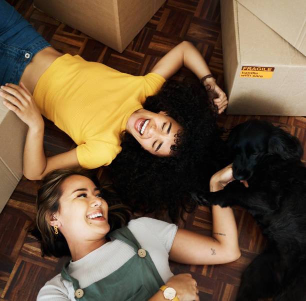 Two friends laughing with a playful dog during a joyful moving day.