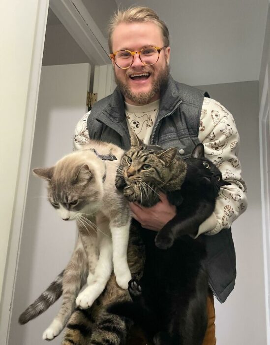 Happy man holding three cats indoors, showcasing love for pets and a joyful moment.