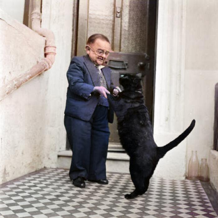 Man in suit playing with black cat on checkered floor near doorway.