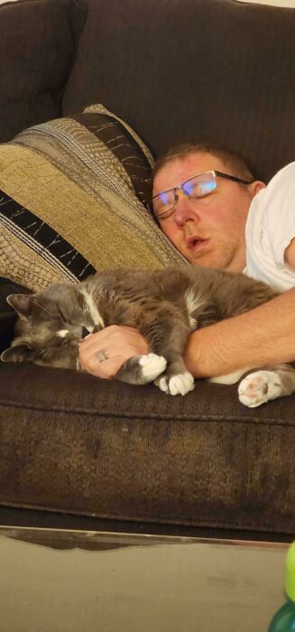 Man and gray cat napping together on cozy brown couch, peaceful home relaxation moment.