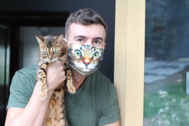 Young man with Bengal cat wearing humorous cat face mask at door.