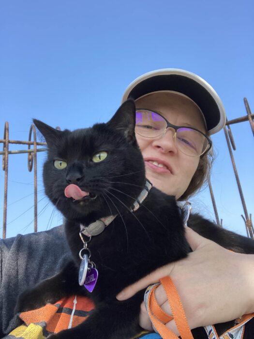 Woman holding black cat outdoors with playful expression and clear sky background.
