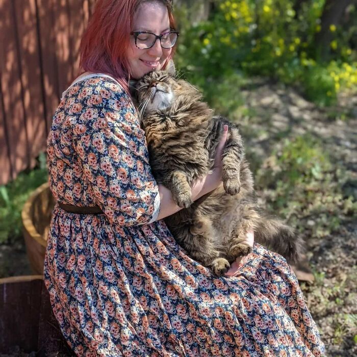 Woman holding fluffy Maine Coon cat outdoors in sunny garden, smiling with joy.