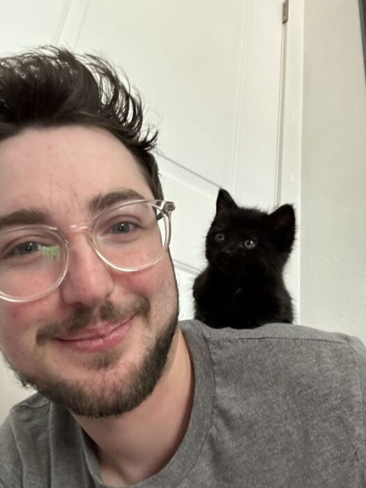 Young man smiling with black kitten on shoulder in cozy indoor setting.
